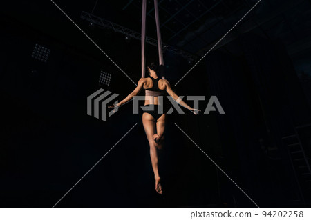 Young woman performing acrobatic element on aerial silk indoors 94202258