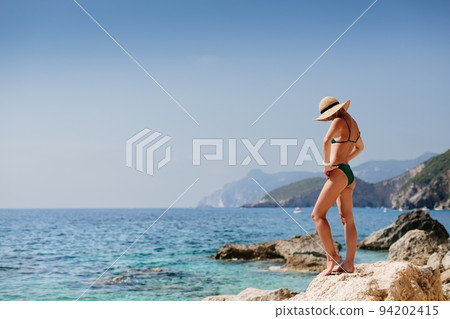 Beautiful blond woman in a green bikini with a straw hat posing on the beach in Corfu Greece.  94202415