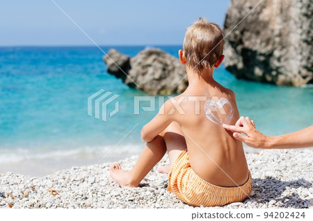 Mom smears her baby's back with sunscreen on a beach in Corfu, Greece. She draws a heart of cream on her son's back.  94202424