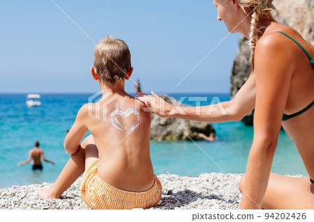Mom smears her baby's back with sunscreen on a beach in Corfu, Greece. She draws a heart of cream on her son's back.  94202426
