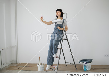 Red hair girl with cute smile holds a paint roller and smart phone in her hands. 94202971