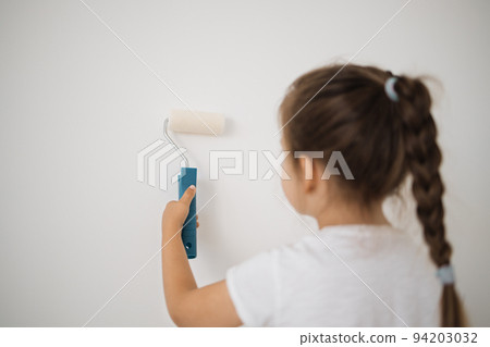 Back view of cute little toddler girl painting the wall with white color in new house. 94203032