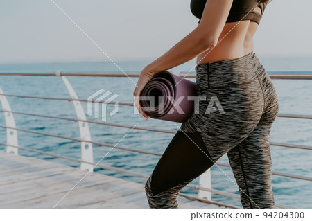 Young sexy sports woman walking to do morning stretching practice. Girl holding yoga mat. Sea or 94204300