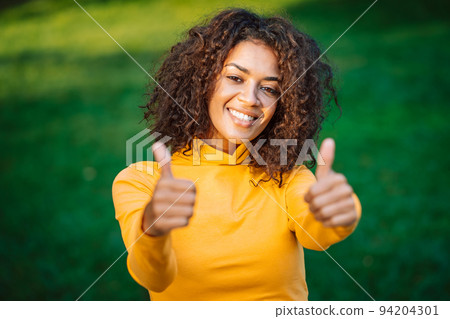Portrait of african american girl showing thumbs up like gesture in green park. Young woman with 94204301
