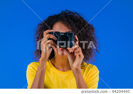 Young african american woman takes pictures with DSLR camera on blue background in studio. Girl Young african american woman takes pictures with DSLR camera on blue background in studio. Girl 94204495