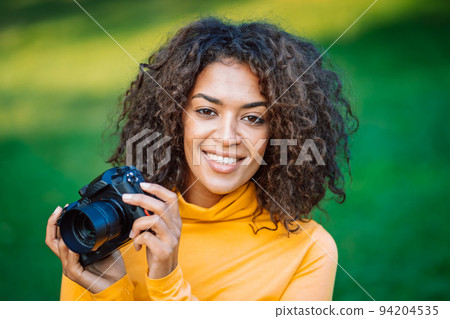 Young pretty african woman in yellow takes pictures with DSLR camera over green background. Girl 94204535