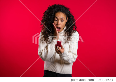 Amazed african american young woman holding small jewelry box with proposal diamond ring on red wall 94204625