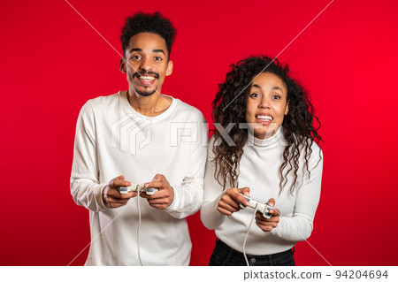 Pretty young african match playing video exciting game on Tv with joystick on red studio wall. Using 94204694
