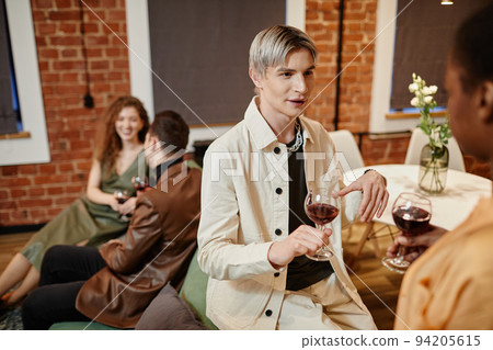 Young gay man in white casualwear holding glass of red wine and talking to girl 94205615