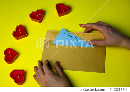 A woman's hand puts a blue note in a cardboard envelope with a handwritten text I LOVE YOU for her lover on Valentine's Day. Love letter on a yellow background flatly. Red candles in shape of hearts. A woman's hand puts a blue note in a cardboard envelope with a handwritten text I LOVE YOU for her lover on Valentine's Day. Love letter on a yellow background flatly. Red candles in shape of hearts. 94205894