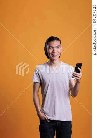 Young smiling asian man using smartphone, messaging on social media. Joyful teenager standing with happy facial expression, holding mobile phone, online communication concept Young smiling asian man using smartphone, messaging on social media. Joyful teenager standing with happy facial expression, holding mobile phone, online communication concept 94207529