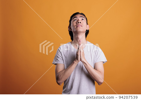 Young man praying folding hands, faith, worship concept. Religious asian teenager pleading, looking upwards with hope, person praising gods, medium shot on orange background Young man praying folding hands, faith, worship concept. Religious asian teenager pleading, looking upwards with hope, person praising gods, medium shot on orange background 94207539