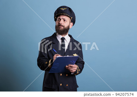 Airplane captain holding clipboard, filling documents, concentrated aviator with form. Aircraft pilot in aviation uniform writing in airport front view, studio medium shot on blue background Airplane captain holding clipboard, filling documents, concentrated aviator with form. Aircraft pilot in aviation uniform writing in airport front view, studio medium shot on blue background 94207574