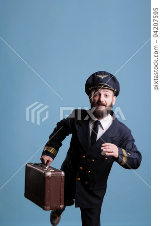 Airplane captain in professional airline uniform running late for flight, carrying vintage suitcase, front view. Pilot with luggage walking in airport, aviator holding baggage, looking at camera Airplane captain in professional airline uniform running late for flight, carrying vintage suitcase, front view. Pilot with luggage walking in airport, aviator holding baggage, looking at camera 94207595