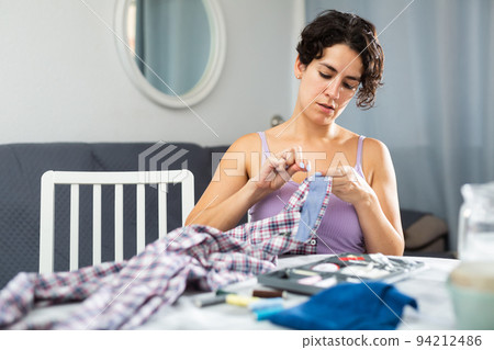 Portrait of attractive focused female using needle and thread to sew shirt Portrait of attractive focused female using needle and thread to sew shirt 94212486