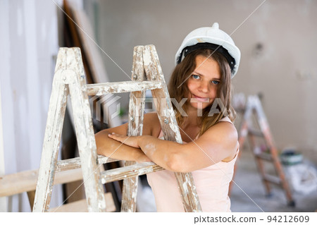 Young attractive girl in beige dress and protective helmet poses on stepladder in room being renovated 94212609