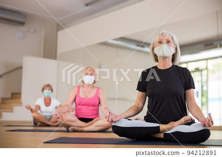 Group of aged women in protective masks, perform an exercise while sitting in the half-lotus position 94212891
