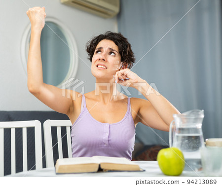 Displeased stressed woman suffering from noisy neighbors from above, covering his ears and shaking fist looking up while trying to reading book 94213089