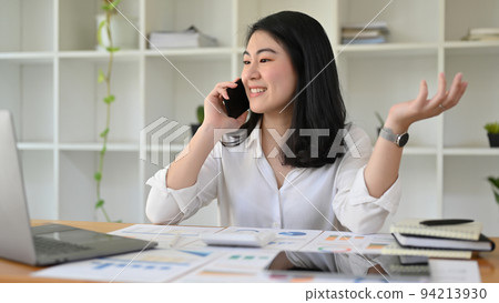 Successful Asian businesswoman at her office desk talking on the phone with her partner. Successful Asian businesswoman at her office desk talking on the phone with her partner. 94213930