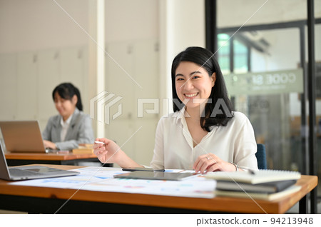 Charming Asian female employee sits at her office desk, working on her financial reports. Charming Asian female employee sits at her office desk, working on her financial reports. 94213948