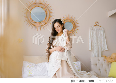 A pregnant young woman has thrown a blanket over her shoulders and is sitting on the bed waiting for the baby 94224888