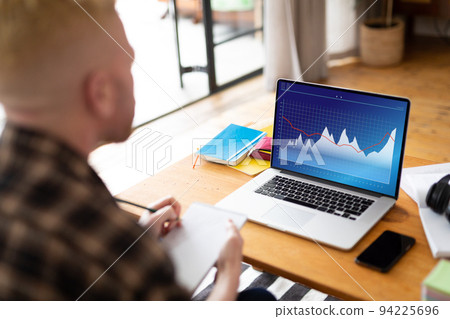 Over shoulder view of caucasian businessman writing notes and using laptop with graphs screen Over shoulder view of caucasian businessman writing notes and using laptop with graphs screen 94225696