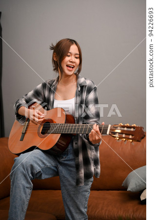 A talented Asian woman sings while playing her acoustic guitar in the living room. 94226693