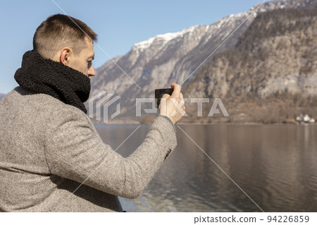 Young adult man taking picture with his smartphone, enjoying mountains, lake, good weather, blue sky, sun. Beautiful, amazing landscape. Tourism, holiday, vacation, travel time. Tourist making photo. Young adult man taking picture with his smartphone, enjoying mountains, lake, good weather, blue sky, sun. Beautiful, amazing landscape. Tourism, holiday, vacation, travel time. Tourist making photo. 94226859