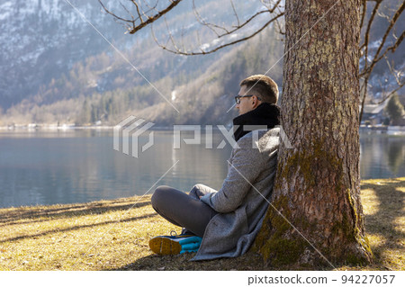 Young man sitting outdoors and enjoying mountains, snow, lake, good weather, blue sky, sun. Beautiful landscape. Time with yourself, dreaming, relaxation, mental health. Tourism, holiday and travel. Young man sitting outdoors and enjoying mountains, snow, lake, good weather, blue sky, sun. Beautiful landscape. Time with yourself, dreaming, relaxation, mental health. Tourism, holiday and travel. 94227057