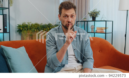 Young man presses finger to lips makes silence hush sign do not tells gossip secret, quiet at home Young man presses finger to lips makes silence hush sign do not tells gossip secret, quiet at home 94229573