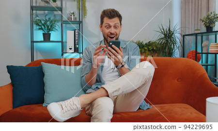 Happy handsome man at home couch use smartphone typing browsing say wow yes found out great big win Happy handsome man at home couch use smartphone typing browsing say wow yes found out great big win 94229695