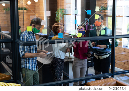 Diverse group of colleagues wearing face masks brainstorming at glass wall in office 94230384