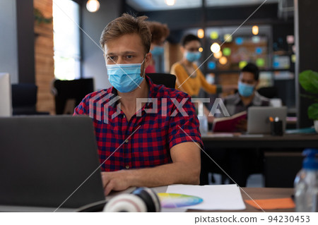 Caucasian man wearing face mask using laptop at desk in casual office Caucasian man wearing face mask using laptop at desk in casual office 94230453