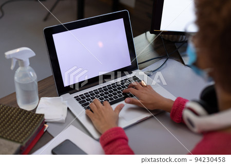Mixed race woman wearing face mask using laptop at desk in casual office Mixed race woman wearing face mask using laptop at desk in casual office 94230458