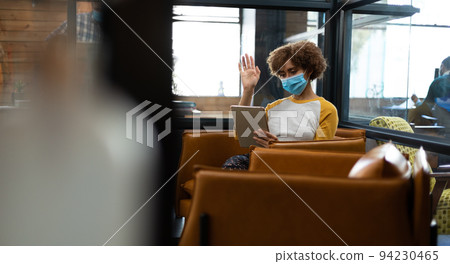 Mixed race woman wearing face mask using tablet and earphones waving during video meeting Mixed race woman wearing face mask using tablet and earphones waving during video meeting 94230465