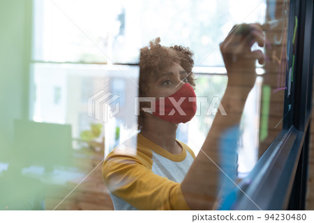 Mixed race woman wearing face mask drawing chart on glass wall in office 94230480
