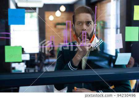 Mixed race man wearing face mask looking at information on glass wall in office Mixed race man wearing face mask looking at information on glass wall in office 94230496