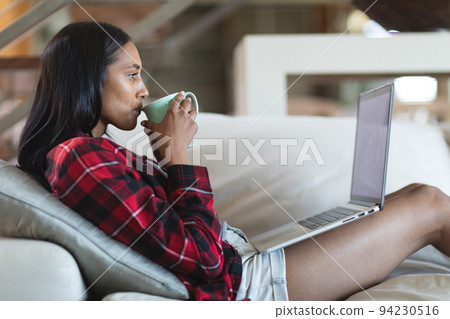Mixed race woman drinking coffee using laptop computer at home 94230516
