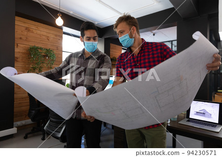 Diverse male colleagues wearing masks in disucssion holding architectural plans in office Diverse male colleagues wearing masks in disucssion holding architectural plans in office 94230571