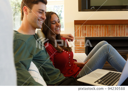 Caucasian couple having video call smiling to laptop sitting in living room Caucasian couple having video call smiling to laptop sitting in living room 94230588