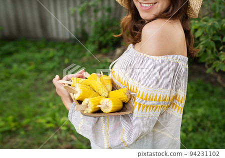 The girl is smiling in a straw hat and holding a plate with boiled corn in her hands. The concept of outdoor recreation, barbecue. The girl is smiling in a straw hat and holding a plate with boiled corn in her hands. The concept of outdoor recreation, barbecue. 94231102