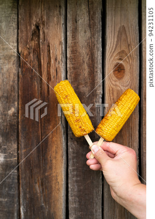In his hands, a man holds fried sweet corn with salt and butter on a stick on a grill against the background of a wooden old wall. Summer vegan dinner or snack, grill cooking. Vertical photo. 94231105