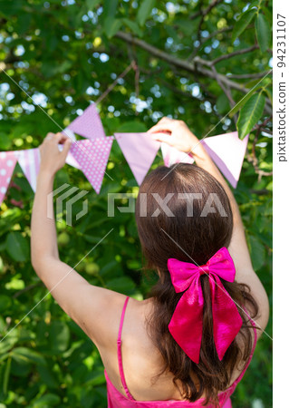 The girl hangs triangular flags of pink color against the backdrop of a green garden. A girl in a pink dress and a bow on her hair. Festive decor - celebration concept. 94231107
