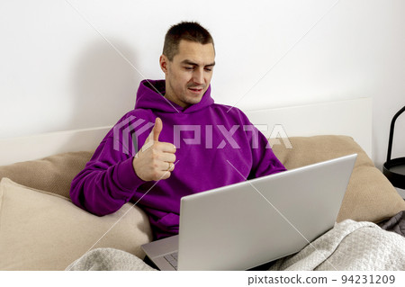 Young caucasian man with violet hoodie sitting on bed and holding laptop computer. Man using notebook to study online, call friends. Distance learning, videocall. Positive sign. Young caucasian man with violet hoodie sitting on bed and holding laptop computer. Man using notebook to study online, call friends. Distance learning, videocall. Positive sign. 94231209