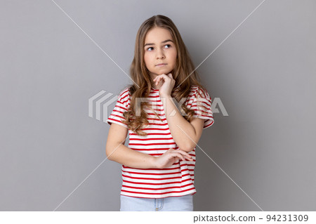 Portrait of thoughtful pensive little dark haired girl wearing striped T-shirt standing holding chin, thinks about something important. Indoor studio shot isolated on gray background. 94231309