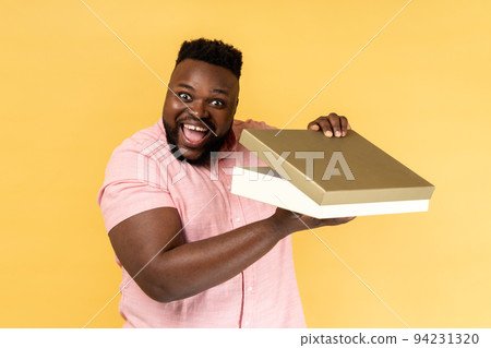Portrait of cheerful bearded man wearing pink shirt opening gift box and looking at camera with toothy smile, satisfied with present. Indoor studio shot isolated on yellow background. 94231320