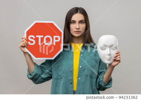 Portrait of serious dark haired woman holding red stop sign and white mask, looking at camera with strict expression, wearing casual style jacket. Indoor studio shot isolated on gray background. 94231362