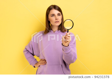 Curious woman looking away through magnifying glass, spying, finding out something, exploring crime scene, inspecting, wearing purple hoodie. Indoor studio shot isolated on yellow background. 94231378