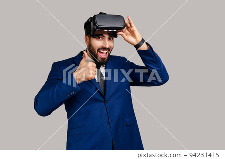 Bearded man with virtual reality glasses expressing happiness after playing game simulator, showing with thumbs up, wearing official style suit. Indoor studio shot isolated on gray background. Bearded man with virtual reality glasses expressing happiness after playing game simulator, showing with thumbs up, wearing official style suit. Indoor studio shot isolated on gray background. 94231415