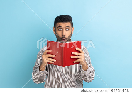 Young adult shocked astonished businessman reading book, reading with big eyes, standing absorbed with amazed plot, wearing striped shirt. Indoor studio shot isolated on blue background. 94231438